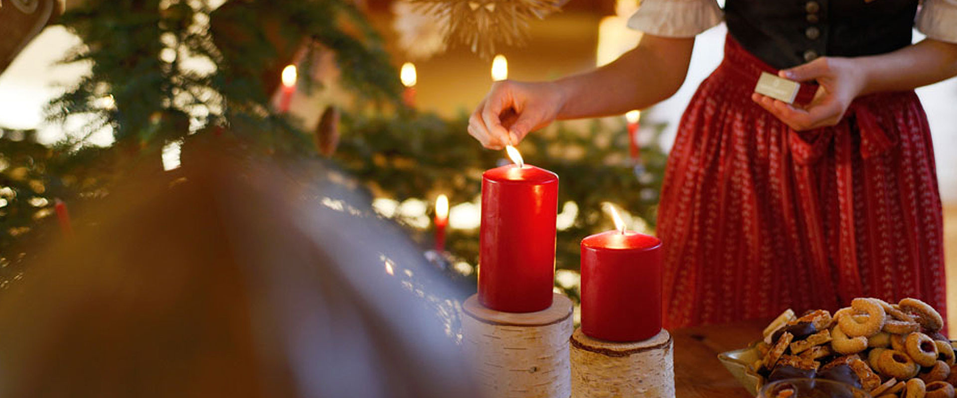 Frau im Dirndl zündet rote Kerze an und daneben sind Weihnachtskekse